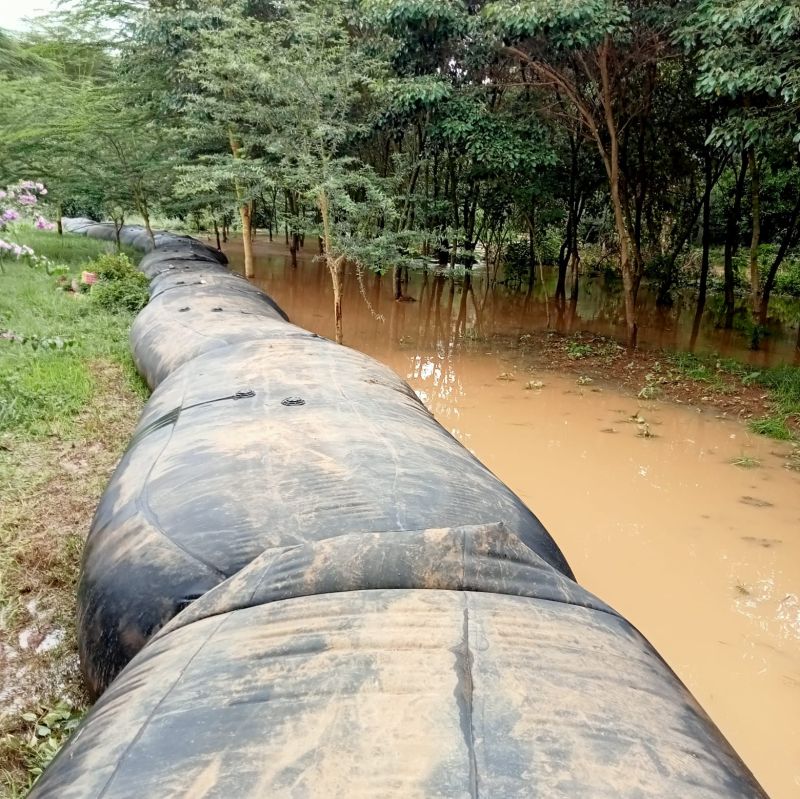 A large black water-filled barrier is set up next to a flooded area near trees and muddy water.