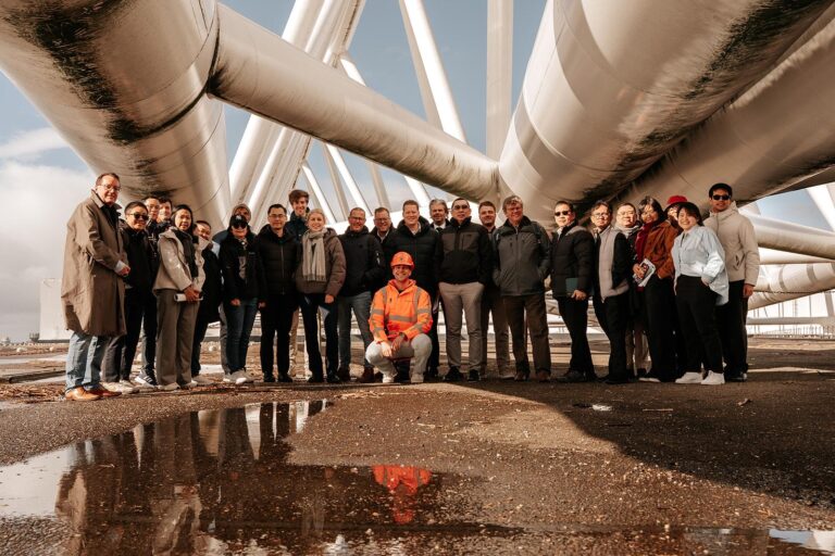 A Thai delegation visits as a group of people stands under large, intersecting metal structures outdoors. One person in an orange safety outfit kneels in front, possibly inspecting the area near a pumping station. The ground is wet with puddles.