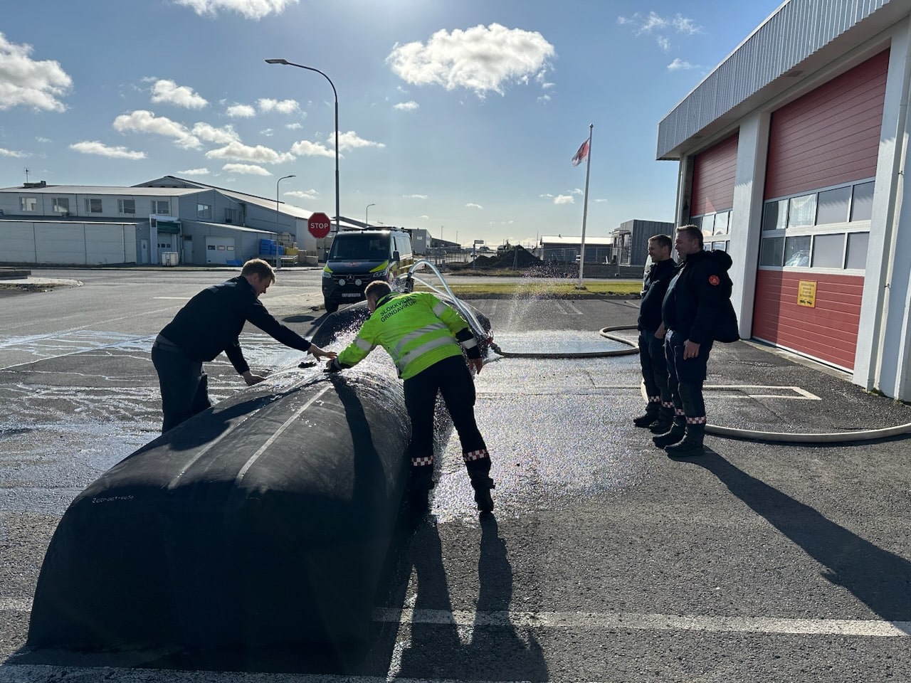 People work with a large, mobile flood barrier outside a building with red doors. A truck and hoses are visible, with some individuals observing.