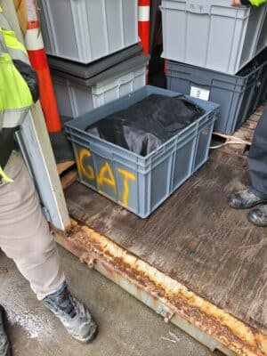 Gray plastic container labeled "CAT" on the side, placed on a wooden platform with other similar containers nearby. People wearing work boots and high-visibility clothing are partially visible around it.