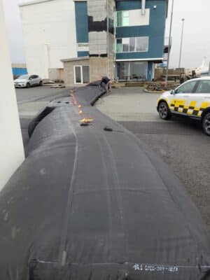 Large black inflatable barrier laid out on a street near a building and parked vehicles, with a person attending to it.