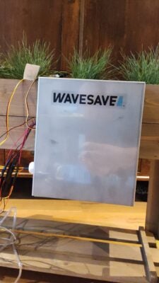 A white box with "WAVESAVE" written on it, attached to wires. It's positioned on a wooden surface with green plants in the background.