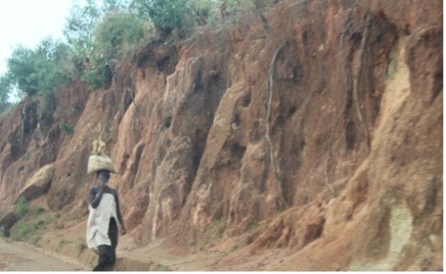 Person carrying items on their head walks along a dirt road with a steep, eroded cliffside in the background.