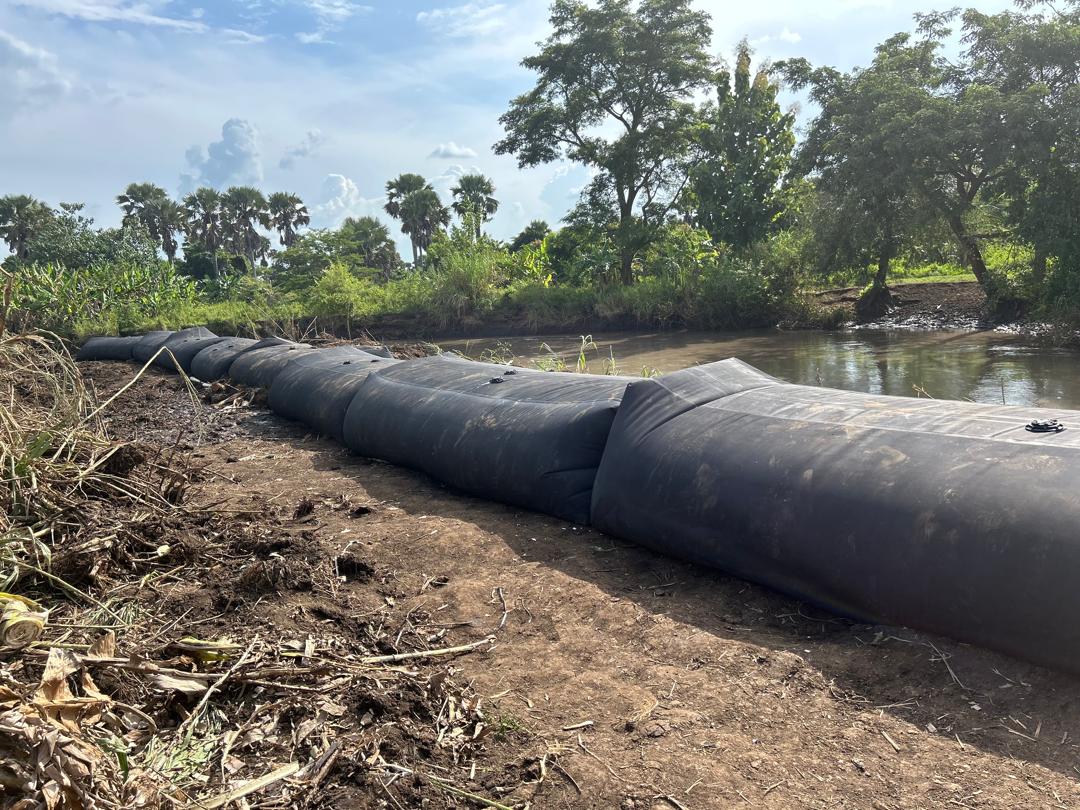Large black sandbags, part of a mobile flood barrier system, are strategically placed along the riverbank with trees and vegetation in the background under a partly cloudy sky.