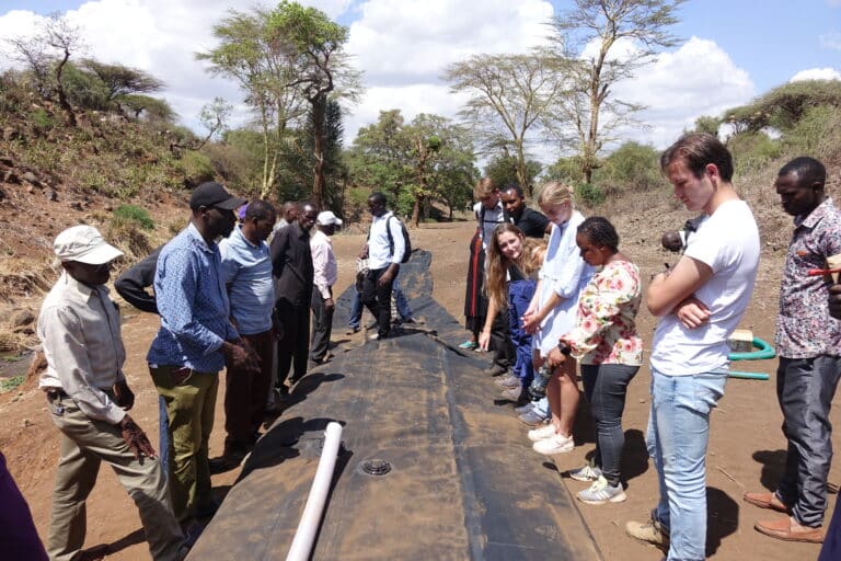 A group of people stand around a large black tarp structure outdoors, discussing or observing it, with trees and cloudy sky in the background.