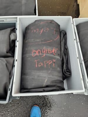 A black tarp labeled "Onýtur Tappi" in red chalk lies in a gray plastic container on a wet ground. A blue shoe is partially visible at the bottom.