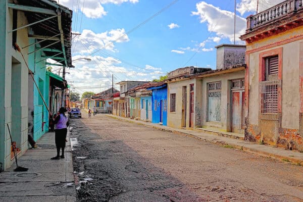 A person sweeps the sidewalk on a quiet street lined with colorful, weathered buildings under a blue sky with scattered clouds.