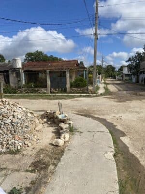 A rural street with houses, power lines, and a pile of rubble or stones on the left. The sky is partially cloudy.