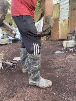 Person in muddy boots, dark pants, and red shirt holds a piece of wood, standing on dirt near corrugated metal structures and debris.