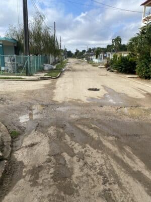 Rural dirt road with potholes and puddles, lined with residential houses and greenery under a cloudy sky.