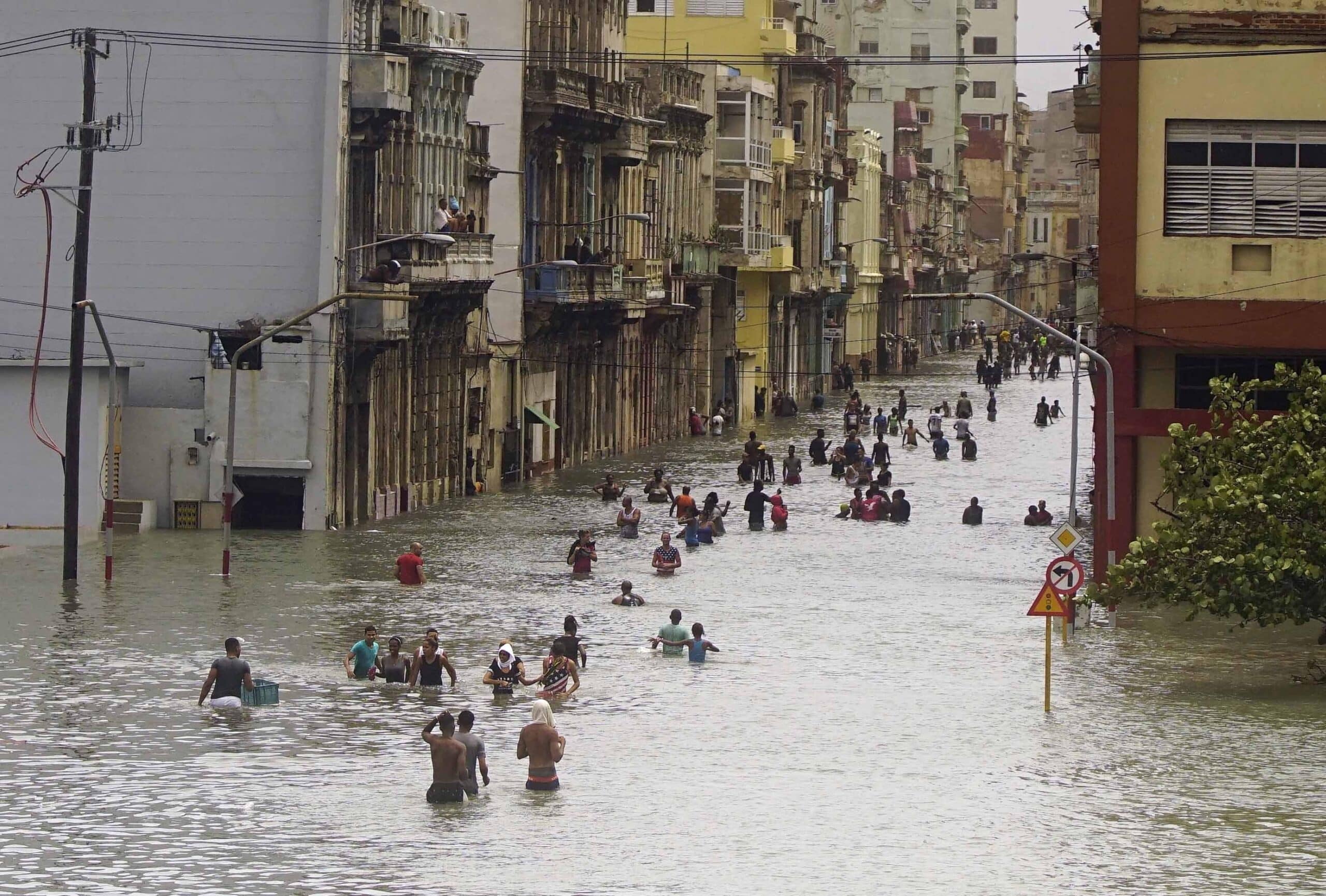 People wading through a flooded street between tall, weathered buildings. Some carry belongings; water reaches knee level. Overcast sky and muted colors indicate a recent heavy rainfall or storm.