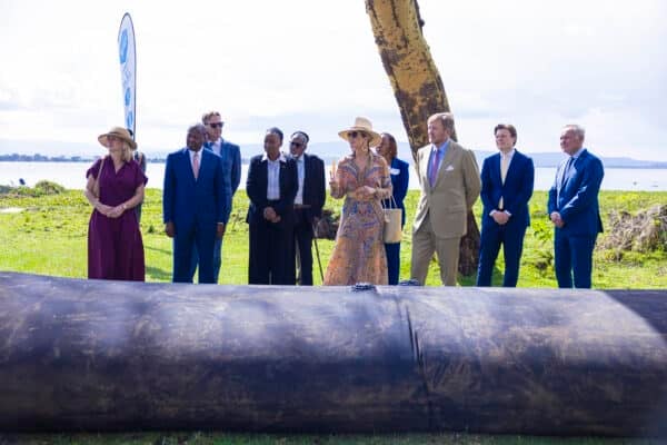 A group of people stands outdoors on grass near a large black pipe, with water and a tree in the background.
