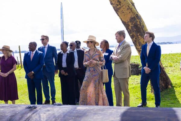 A group of people, including men in suits and a woman in a patterned dress and hat, stand outdoors near a tree, with a lake and grassy area in the background.