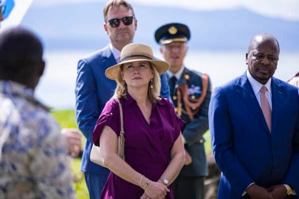 A group of people, including a woman in a hat and a man in a blue suit, stand outdoors with a lake and mountains in the background. Two other men, one in uniform, stand behind them.