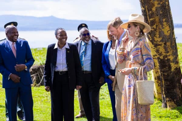 A group of formally dressed people stand on grass near a tree and a body of water, smiling and talking on a sunny day.