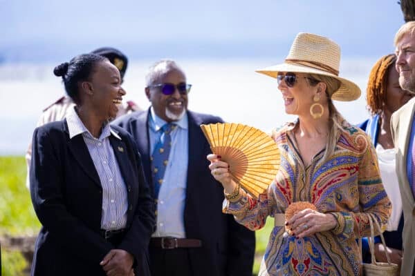 A group of people stand outdoors, conversing and smiling; one woman in a hat and sunglasses holds a handheld fan.