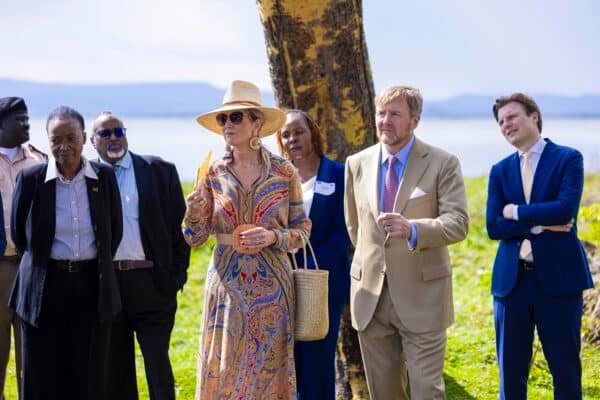 A group of people, including a woman in a patterned dress and sunhat and a man in a light suit, stand outdoors near a tree, listening attentively.