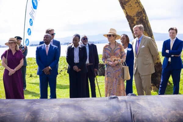 A group of people, some in suits and formal attire, stand outdoors on grass near a large tree, with a body of water and hills in the background.
