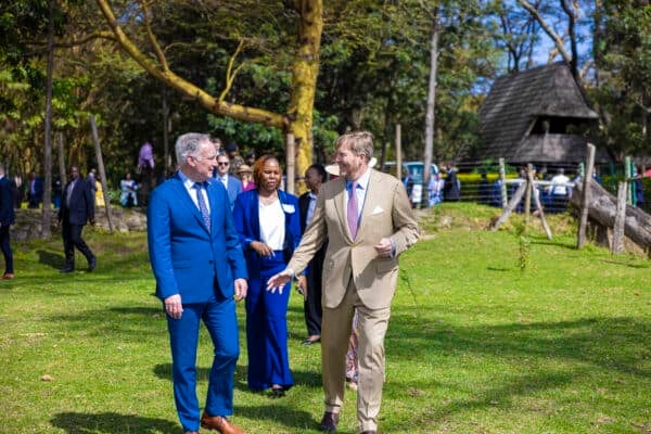 Two men in suits walk and talk on a grassy area, with a woman and other people in the background near trees and a fence.