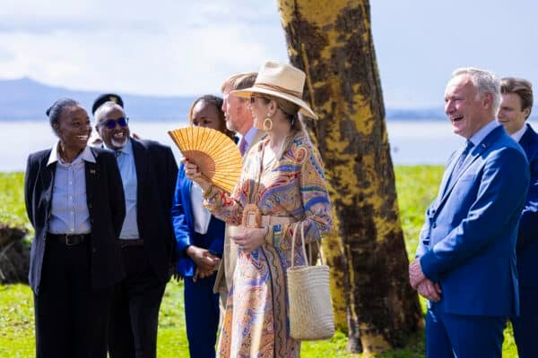 A group of people, some in suits, stand outdoors on grass near a tree; a woman in a hat holds a folding fan and smiles while others look on.