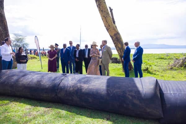 A group of people, some in formal attire, stand on grass near a large black tube with a lake and trees in the background.