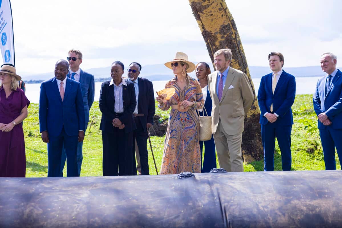 A group of men and women stand outdoors in formal attire, some wearing suits and hats, near a large black pipe during a WaveDam mobile flood barrier demonstration, with greenery and water in the background.