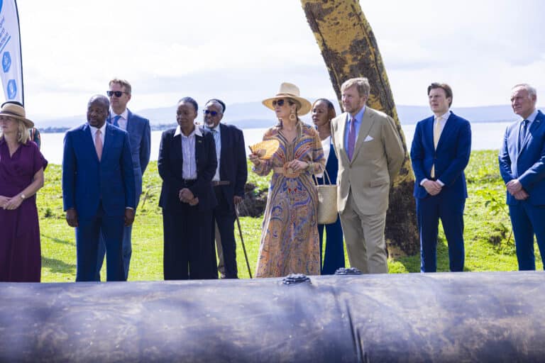 A group of men and women stand outdoors in formal attire, some wearing suits and hats, near a large black pipe during a SLAMDAM mobile flood barrier demonstration, with greenery and water in the background.