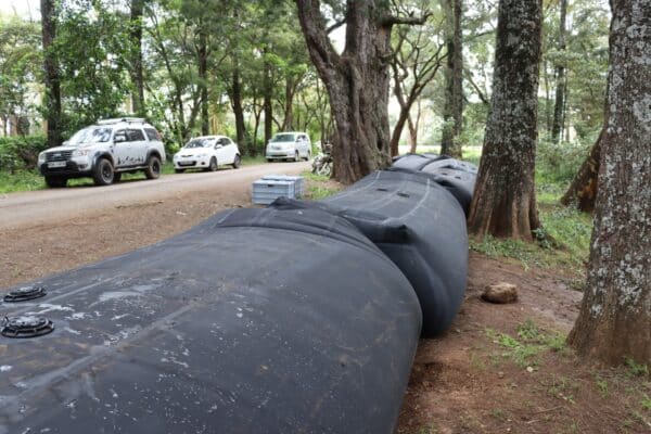 Large black plastic water tanks lie on the ground beside a dirt road with several parked cars and trees in the background.