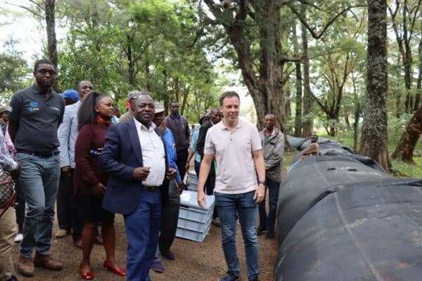 A group of people stand outdoors near large cylindrical black tanks, surrounded by trees, with some individuals engaged in conversation.