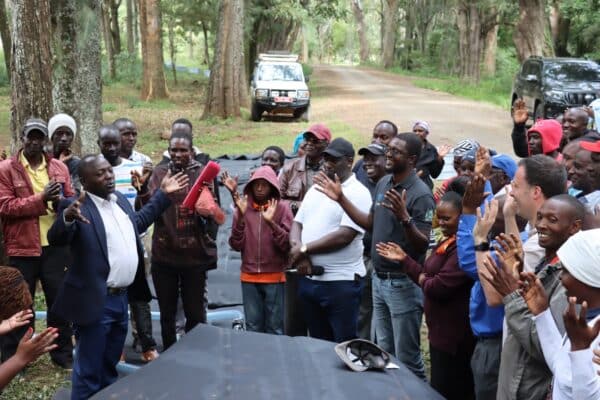 A group of people stand outdoors in a wooded area, some clapping and others listening, with two vehicles parked on a dirt road in the background.