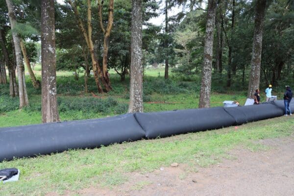 A long black inflatable barrier is set up along the edge of a wooded area, with several people standing nearby.
