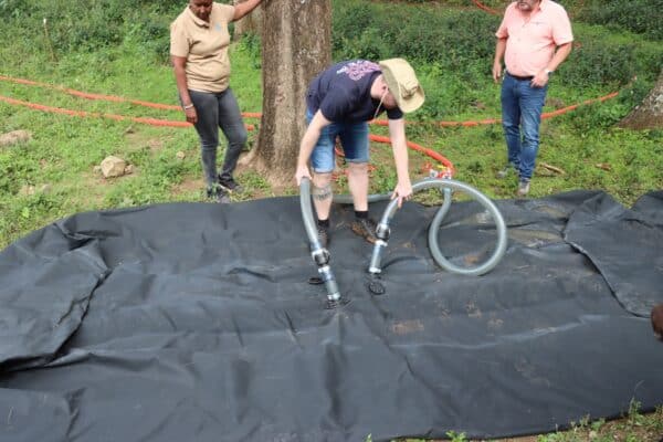 Three people stand on grass as one places two large hoses onto a black tarp spread on the ground; red hoses and trees are visible in the background.