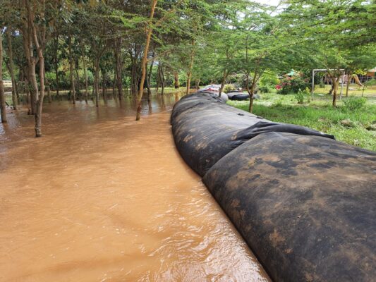 A large black water-filled barrier, used for flood control in Kenya, separates floodwater from a grassy area with trees and some visible playground equipment in the background.