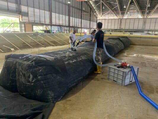 Two people fill a large black water bladder with hoses inside a spacious industrial facility with open walls and a high ceiling.