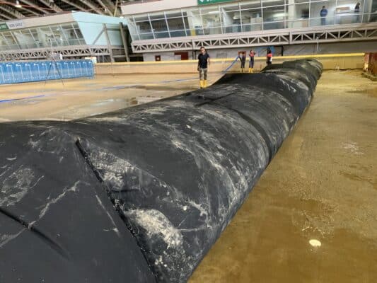 Large black water-filled barrier placed on a wet indoor floor with several people in boots standing nearby, and portable toilets in the background.