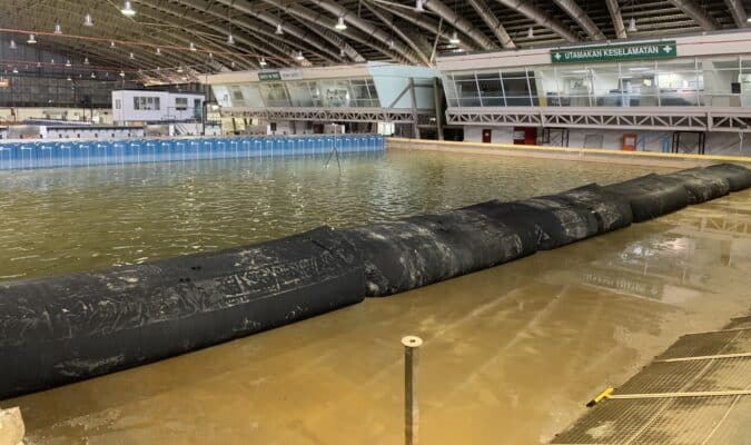 Large indoor pool with a raised barrier dividing the water, situated in a spacious facility with SLAMDAM Certified for Flood Protection signage on the walls and a high arched ceiling.