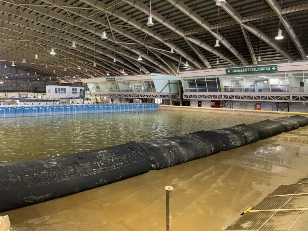 An indoor facility with a large pool of water, temporary barriers, and a sign reading "UTAMAKAN KESELAMATAN," likely indicating a safety warning in Malay.