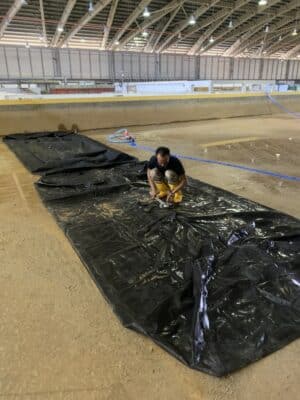 A person kneels on a large black plastic sheet laid out on the ground inside a spacious indoor facility with high ceilings.