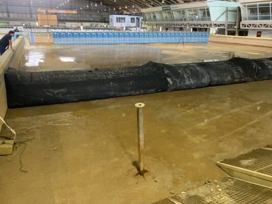 Large indoor pool area under maintenance with water partially drained, exposing muddy floor, black barrier, and blue portable toilets lined up in the background.