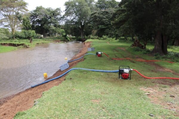 Two portable water pumps with red and blue hoses are set up on a grassy riverbank, drawing water from the river under a cloudy sky.