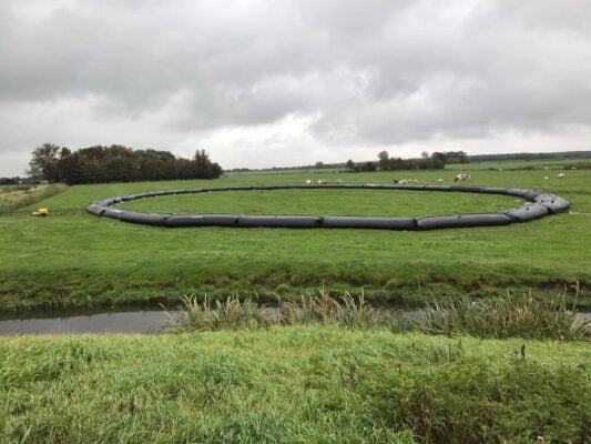 A large black circular barrier is placed on a grassy field with cows grazing in the distance and a small canal in the foreground under a cloudy sky.