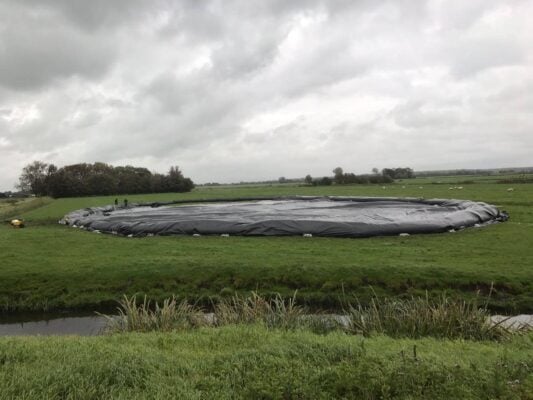 A large black plastic-covered circular structure sits on a grassy field under a cloudy sky, with a ditch and vegetation in the foreground.