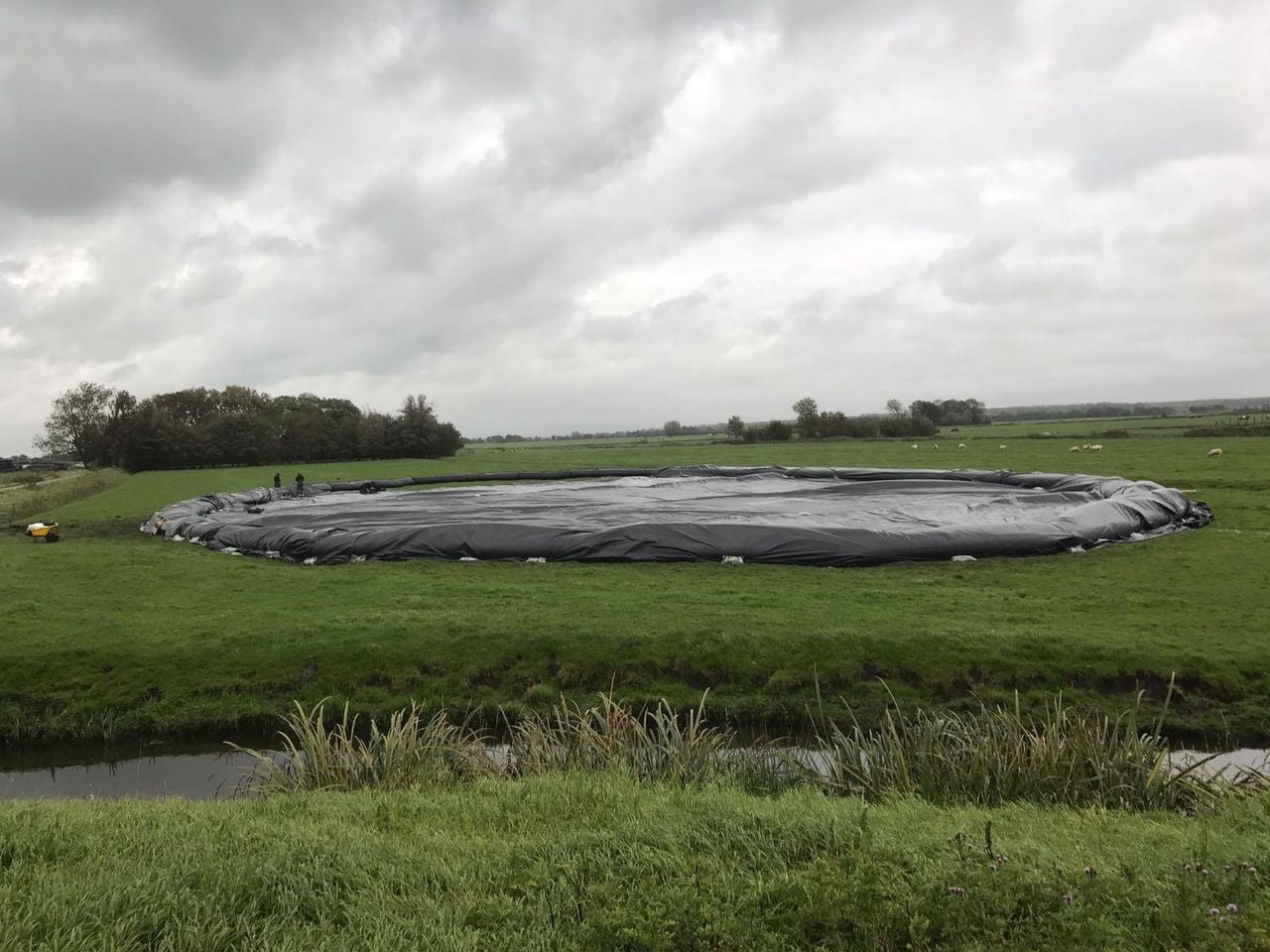 A large black plastic-covered circular structure sits on a grassy field under a cloudy sky, with a ditch and vegetation in the foreground.