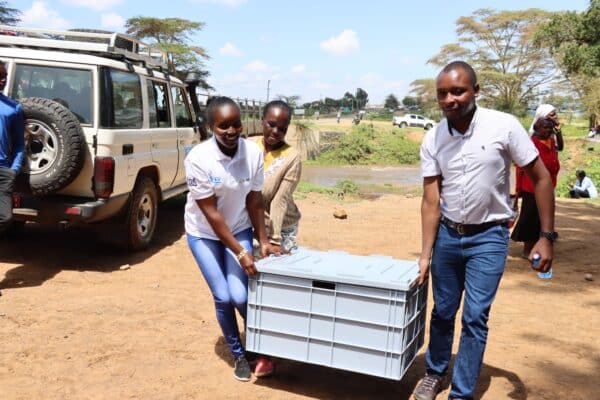 Three people carry a large gray plastic container outdoors near a vehicle, with others and trees visible in the background.
