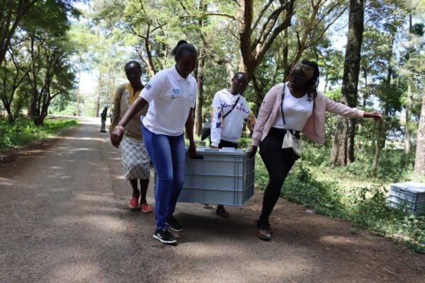 Four people walk along a tree-lined road in Kenya, carrying a large plastic container together—likely part of local flood control efforts; additional containers are visible on the ground nearby.