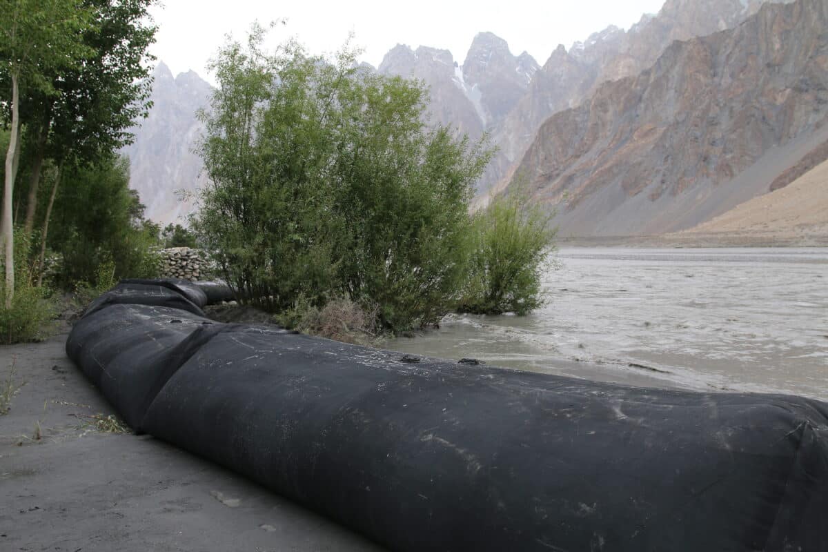 A large black water-filled flood barrier, a Climate Innovation Challenge winner, lies on muddy ground near a river, with trees and mountains in the background.