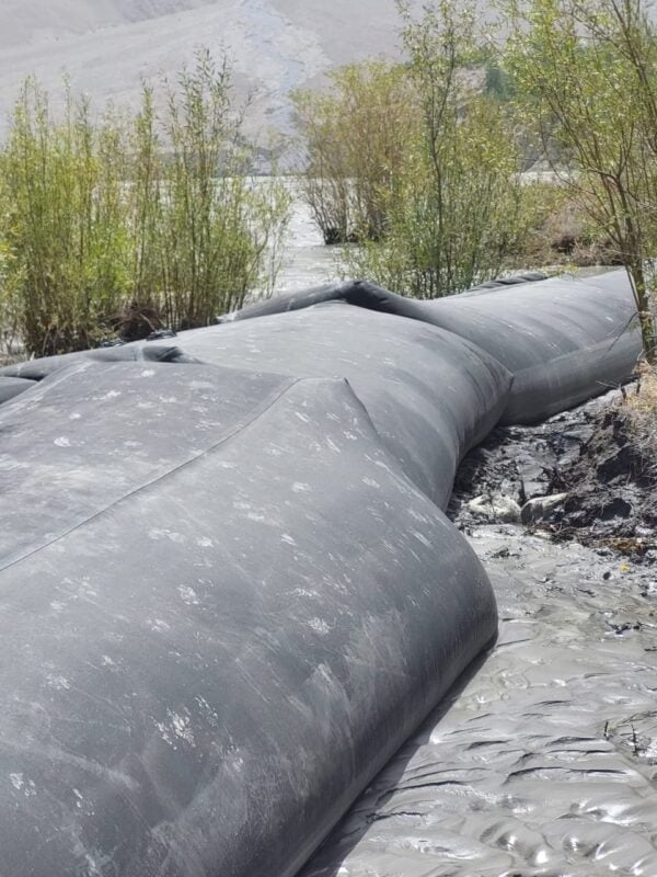 A large black geotextile tube, a Climate Innovation Challenge winner, lies on muddy ground near water and green shrubs, with mountains visible in the background.