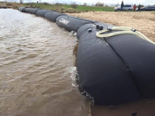 Large black flood barrier tube filled with water lines the shoreline, hoses attached, effectively separating water from sandy land; cars and people are visible in the background.