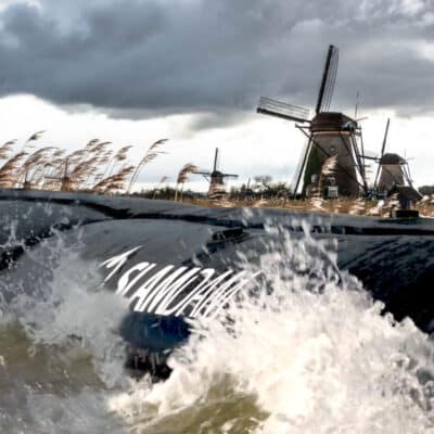 A large wave splashes against a black flood barrier marked "ISLAND," with windmills and tall grass visible in the background under a cloudy sky.