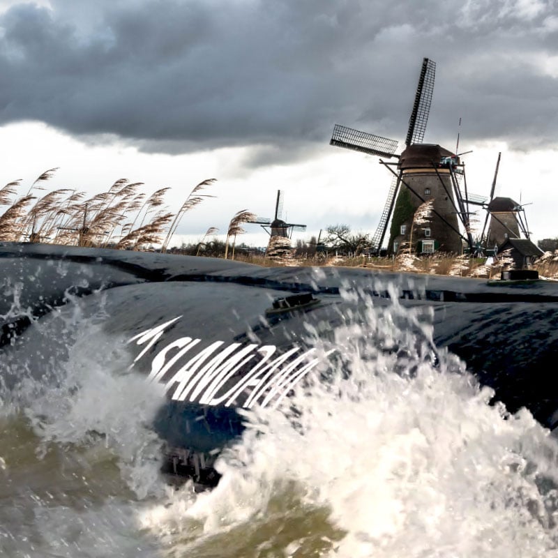 A large wave splashes against a black flood barrier marked "ISLAND," with windmills and tall grass visible in the background under a cloudy sky.